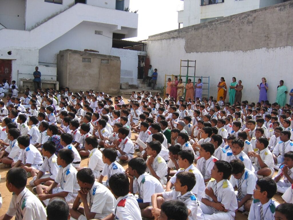 Large group of schoolchildren in uniform seated outdoors during an assembly, with teachers standing along a wall behind them.