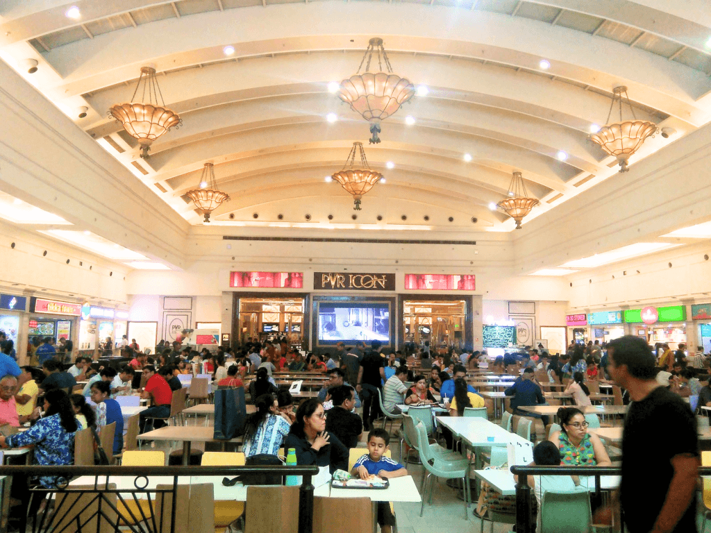 Busy shopping mall food court with diners seated at tables and a cinema entrance in the background.