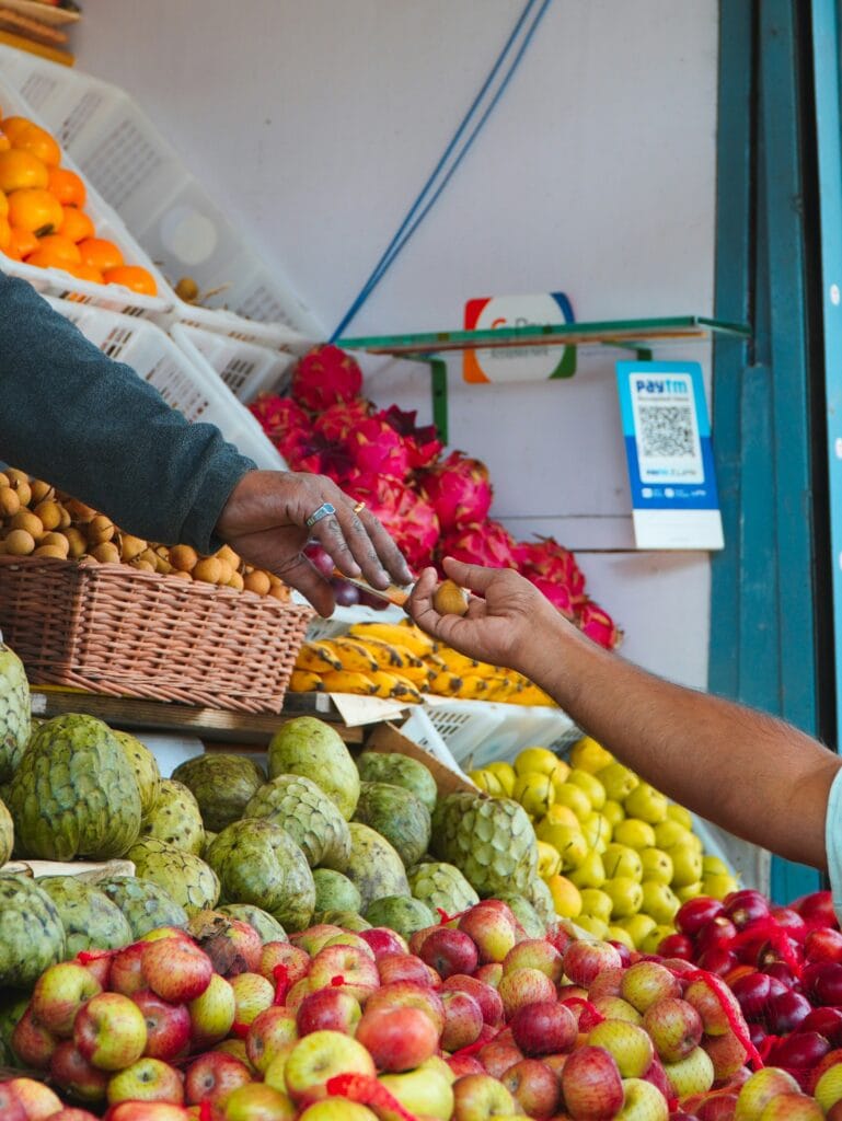 Customer paying a fruit vendor with cash at a market stall, with a digital payment QR code displayed nearby, UPI