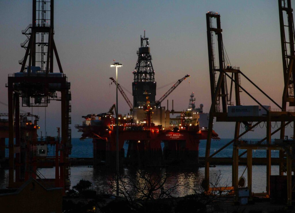 Night view of a semi-submersible offshore oil rig, the West Bollsta, illuminated at a shipping port with large industrial gantry cranes in the foreground.